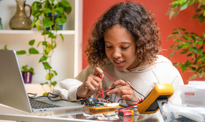Young student working on an electronics project with a multimeter and circuit board at a desk, smiling and focused, with a laptop and components in a brightly colored, plant-filled room.
