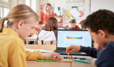 Two students working together on an electronics project at a classroom table, with a laptop displaying a color-coded chart in the background and other children and a teacher visible behind them.
